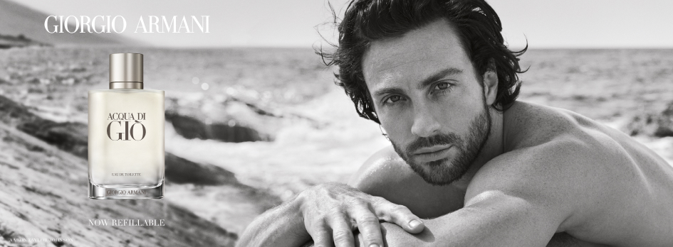 Werbung für Giorgio Armani Acqua di Giò Eau de Toilette. Rechts ist ein muskulöser Mann mit dunklem Haar und Bart, der oberkörperfrei am Felsenstrand liegt und direkt in die Kamera blickt. Links daneben eine Flasche des Parfums mit der Aufschrift 'Acqua di Giò Eau de Toilette Giorgio Armani'. Darüber der Markenname 'Giorgio Armani' und darunter der Text 'Now Refillable'. Die Szene ist in Schwarz-Weiß gehalten.