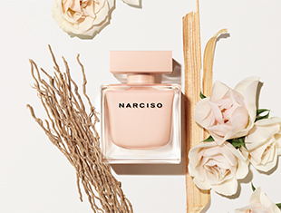 Quadratischer, roséfarbener Parfumflakon mit der schwarzen Aufschrift 'NARCISO' liegt auf hellem Hintergrund. Umgeben ist er von beigen Blüten, zarten Holzstücken und getrockneten Zweigen – symbolisch für die natürlichen Duftnoten des Parfums.